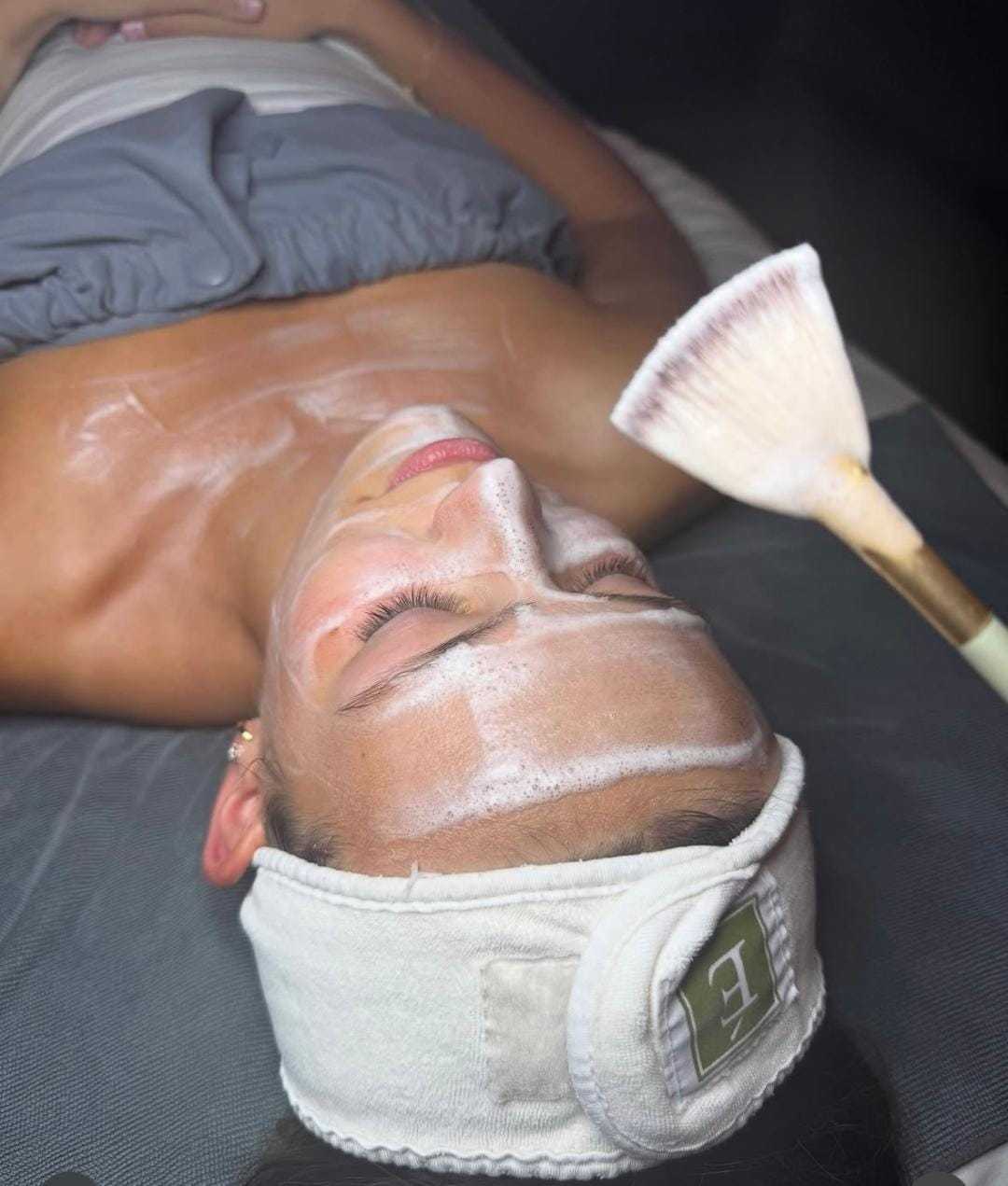 Person receiving a facial treatment with a brush applying white cream on their face.