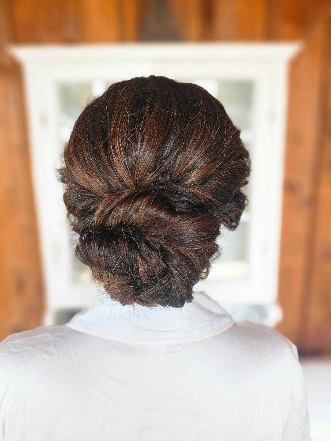 Elegant brunette updo hairstyle against a blurred white and wood background.